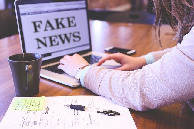 jeune femme écrivant sur son ordinateur portable posé sur une table à côté d'une tasse et d'un bloc de feuilles. Il est écrit sur l'écran "fake news"